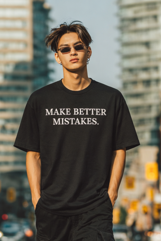 Person wearing a black t-shirt with 'MAKE BETTER MISTAKES' text on a city street.